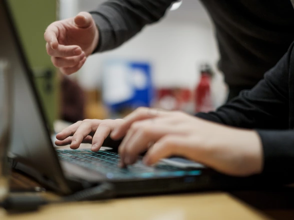 Ein Laptop, an dem zwei Hände arbeiten. Eine weitere Hand zeigt etwas auf dem Bildschirm.